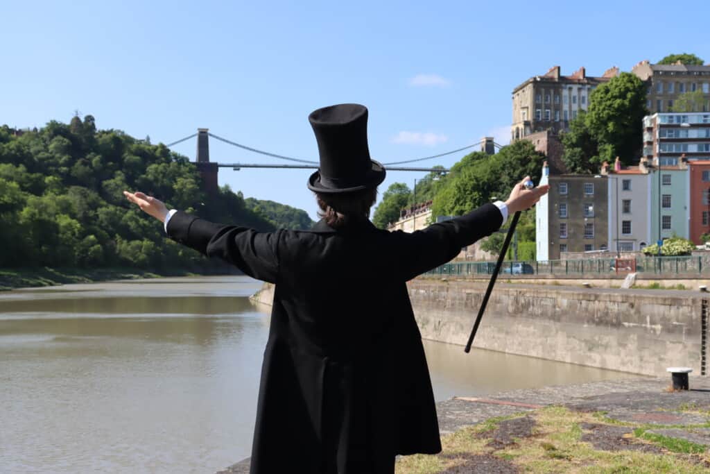 A man in historical clothing is holding his arms out to the side as he looks at a large bridge.