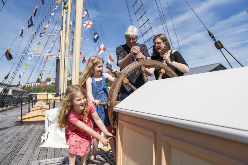 A group of people, two adults and two children, are on board a ship, holding handles of the steering wheel.
