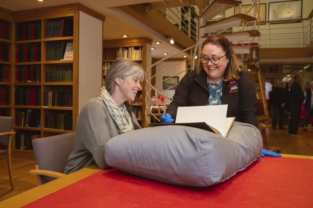 A woman with grey plaited hair sits at a red wooden desk. Another woman stands next to her showing her a book that has been placed on a pillow. They are both smiling. In the background are more people and bookcases filed with leather-bound books.