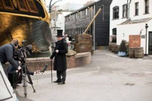 A man dressed as Mr Brunel in a Victorian Style long coat and top hat stands in front of a camera crew who point a sound boom at him. Behind them is a Victorian Steamship the SS Great Britain.