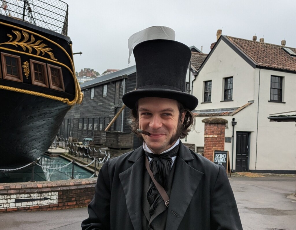 A man dressed as Mr Brunel, a Victorian gentleman in a black suit with waistcoat and jacket. He is smirking and looking to his left. There is a roll of toilet roll on the top of his tall black stovepipe hat.