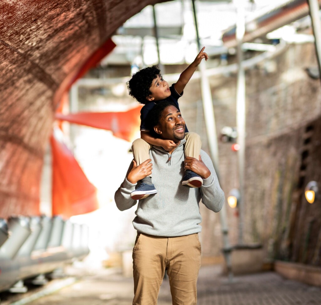 A father carrying his son on his shoulders who is pointing excitedly at something in the dry dock of the SS Great Britain. 