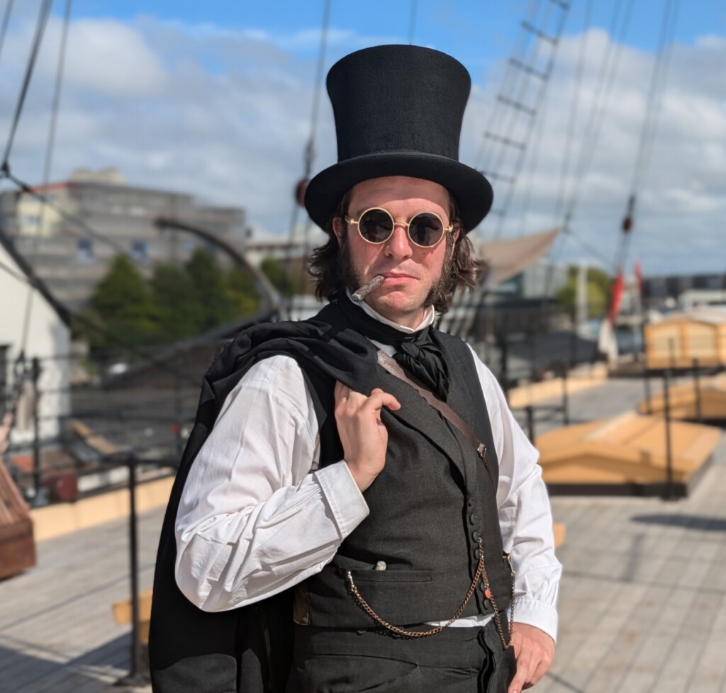 Mr Brunel the Victorian engineer dressed in a black and white suit with top hat and sunglasses poses on the ship's deck 